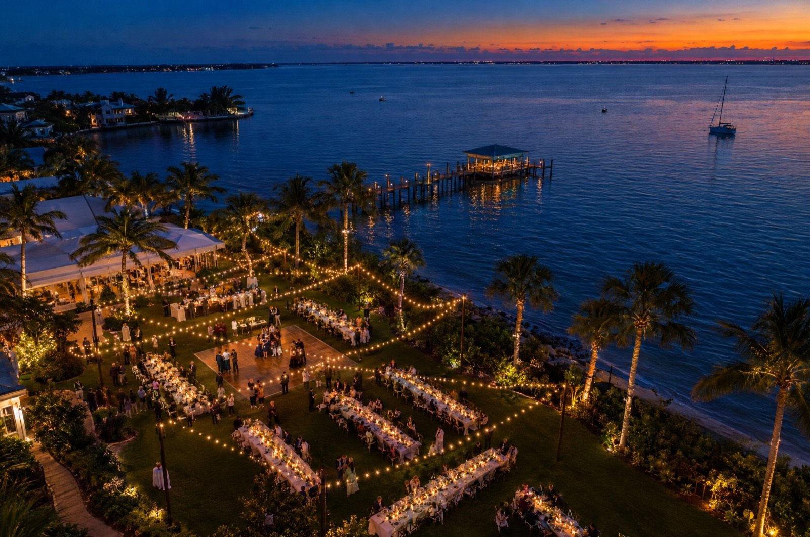 Aerial view of a string-lit waterfront dinner party at dusk